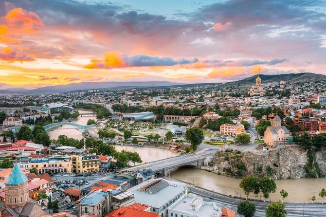 Panoramic view of Tbilisi, Georgia, featuring the Kura River, Peace Bridge, historic Old Town, modern architecture, and the Holy Trinity Cathedral under a colorful sunset sky