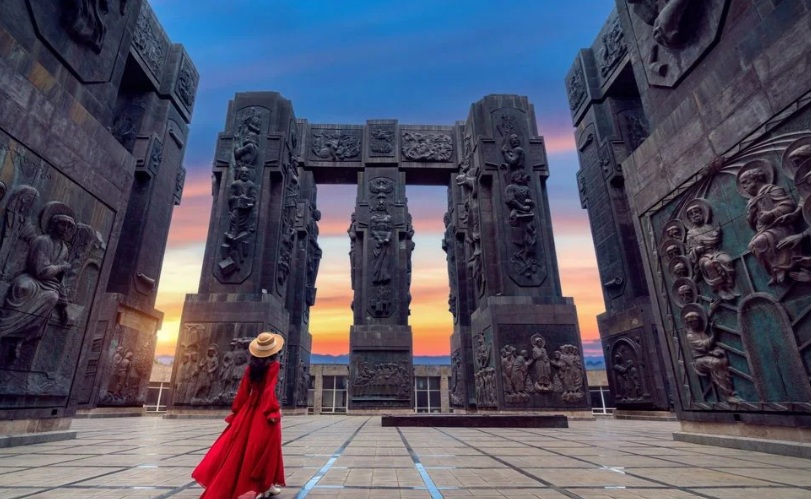 A traveler in a flowing red dress stands before the towering Chronicle of Georgia monument in Tbilisi during sunset, surrounded by massive stone pillars carved with historical and religious reliefs