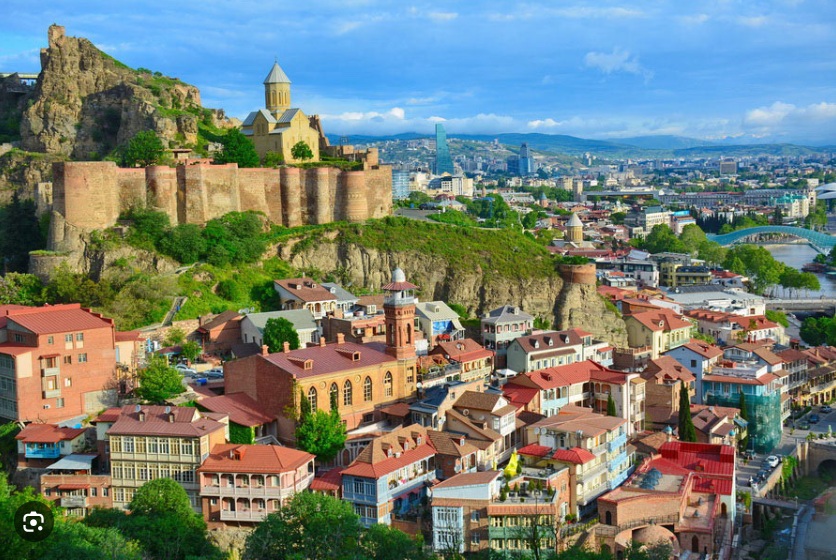 Elevated view of Old Tbilisi with Narikala Fortress, St. Nicholas Church, traditional houses with red roofs, the Kura River, and the city skyline beneath dramatic clouds
